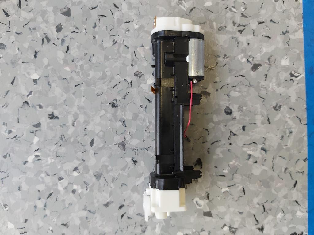 Elektrischer Linearantrieb,
- bestehend aus einem rotierenden Gleichstrommotor für eine Leistung von weniger als 37,5 Watt (3,3 VDC Nennspannung) mit zweipoligen Anschlusskabel, einem optischen Inkrementalgeber mit Flachbandanschlusskabel, einem mehrstufigen Zahnradgetriebe und linear arbeitendem Stellglied (Kolbenstange), montiert an einem mehrteiligen Kunststoffträger mit Aufnahmeschacht für eine Stromversorgungsquelle und Montagevorrichtungen,
- nach Versorgen des Gleichstrommotors mit Nennspannung wird die Kolbenstange je nach Polung ausgefahren oder eingezogen,
- die Hublänge beträgt ca. 68,5 mm.
(Abbildung laut Anlage)
