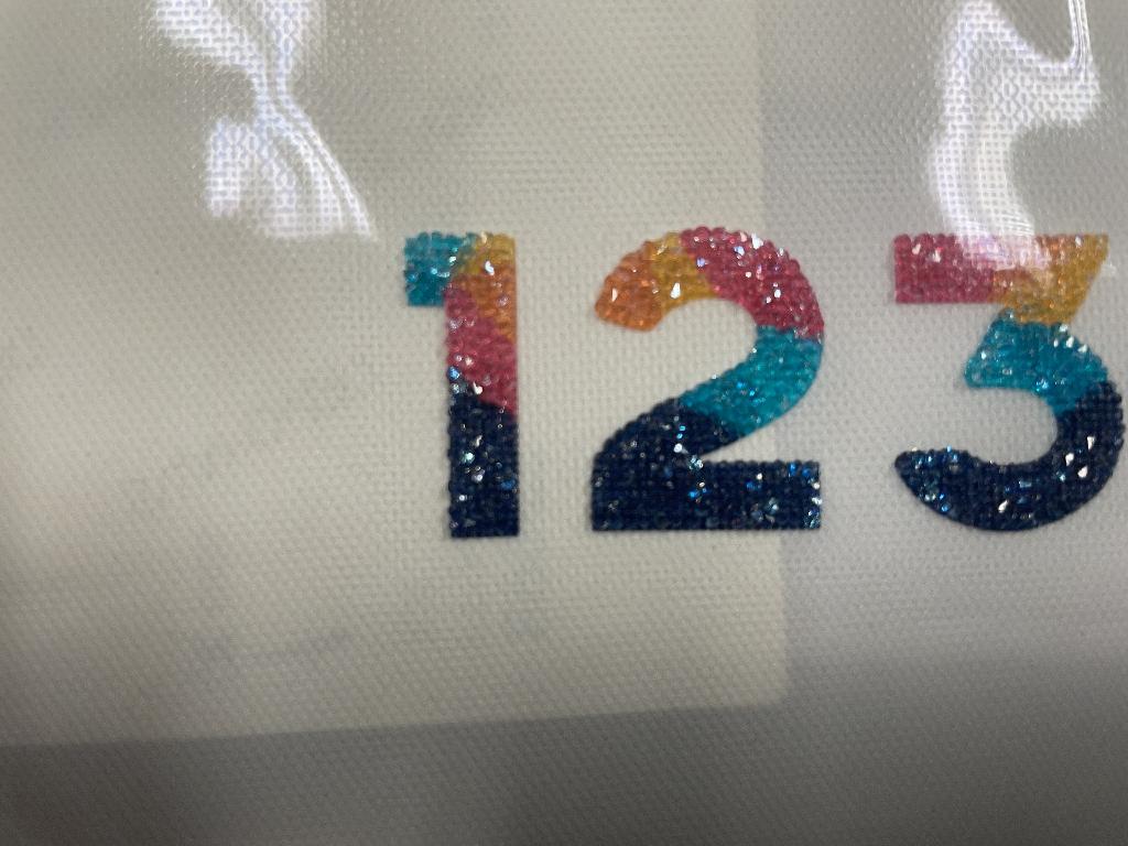 Nachahmungen von Schmucksteinen und ähnliche Glaskurzwaren und Waren daraus
- in Form von Buchstaben/Zahlen, 
- bestehend aus Strasssteinen mit geschliffenen Kanten und Flächen, welche auf selbstklebenden und unterschiedlich eingefärbten Unterlagen aufgebracht sind.
(Abbildung siehe Anlage)