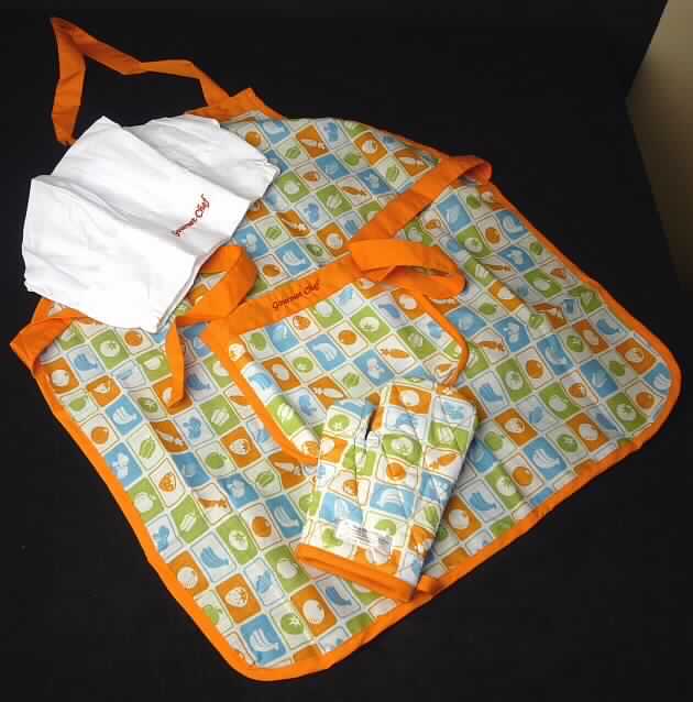 Dreiteilige Warenzusammenstellung, bestehend aus einer anderen Kleidung (Schürze), einer Kopfbedeckung und
einem Fausthandschuh, sog. Spielzeug-Schürze, Mütze und Handschuh, Art.Nr.: 821774 (E3119), Foto siehe Anlage,
- gemeinsam in einem Pappkarton für den Einzelverkauf aufgemacht und zur Befriedigung eines speziellen
  Bedarfs und zur Ausübung einer bestimmten Tätigkeit zusammengestellt,
- laut Verpackung für Kinder ab drei Jahren,
- alle Bestandteile aus laut Verpackung 80 % Polyester (synthetische Chemiefasern) und 20 % Baumwolle,
- andere Kleidung (sog. Spielzeug-Schürze):
-- aus ca. 0,3 mm dicken, buntbedruckten Geweben, 
-- den vorderen Teil des Körpers bedeckend, 
-- in den Abmessungen: ca. 59 cm x 52 cm,
-- vorn mit einer aufgesetzten, offenen Tasche, mit einem aufgestickten Schriftzug "Gourmet Chef" verziert,
-- ohne Merkmale, die auf einen bestimmten Trägerkreis hinweisen (unisex), 
-- mit schmalen Streifen aus Geweben eingefasst,
-- mit Nackenträger und Bindebänder in der Taille aus Geweben,
-- u. a. aufgrund der Größe keine Einreihung als Arbeits- und Berufskleidung in die Unterposition 6211 4210,
-- bestimmt nach dem Gewicht und dem Gesamteindruck den Charakter der Warenzusammenstellung, 
- Kopfbedeckung (sog. Mütze):
-- aus Zuschnitten aus ca. 0,3 mm dicken, einfarbigen Geweben hergestellt, 
-- flachliegend in den Abmessungen: ca. 27 cm breit und 24 cm hoch,
-- ohne Schirm,
-- vorn mit einem aufgestickten Schriftzug "Gourmet Chef" verziert,
-- am unteren Rand mit seitlich eingearbeiteten, elastischen Bändern ausgestattet,
- Fausthandschuh mit Daumen (sog. Handschuh):
-- in den Abmessungen: ca. 20 cm x 12 cm,
-- aus einem dreilagig abgeteilt versteppten Verbunderzeugnis der Position 5811 mit einer Außenlage aus
   dem gleichen buntbedruckten Geweben wie die Schürze, einer Innenlage aus Vliesstoff und einer
   Zwischenlage aus Wattierungsstoff,
-- an der Öffnung mit einem Band aus Geweben eingefasst,
-- mit einer Aufhängeschlaufe.

"Andere Kleidung (Schürze), aus Geweben, für Mädchen, aus Baumwolle, keine Arbeits- und Berufskleidung"
