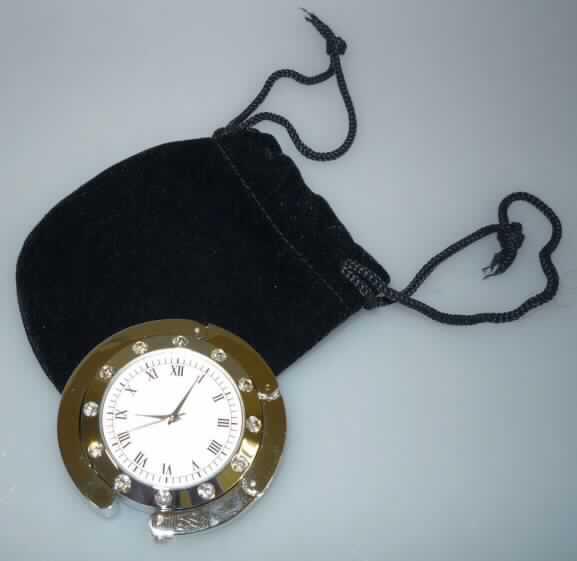 Runde Taschenuhr, sog. Handtachen-Butler, Art.Nr. 8020500,
mit einem batteriebetriebenen Quarzuhrwerk mit mechanischer Anzeige (Stunden, Minuten, Sekunden). Das Gehäuse besteht aus unedlem Metall und einer aufgeklebten rutschfesten Gummiunterseite (Durchmesser ca. 38 mm, Dicke ca. 12 mm) und ist auf der Oberseite mit 12 geschliffenen Glassteinen eingefasst. An der Gehäuseaußenseite befindet sich ein falt- und abklappbarer Metallbügel, an den, wenn die Uhr an eine Tischkante gelegt wird, eine Handtasche angehängt werden kann.

Die Uhr ist in ein verschließbares Veloursäckchen eingelegt und in einer Pappschachtel verpackt.
Das Erzeugnis ist als "Taschenuhr, elektrisch betrieben" einzureihen.