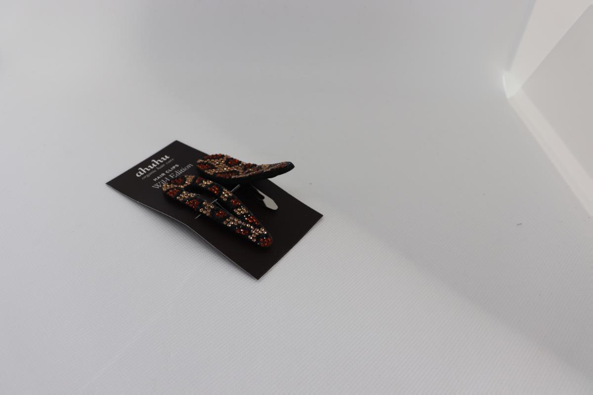 Bei den sog. "Haarspangen aus Kunststoff" handelt es sich um zwei aus verschiedenen Stoffen zusammengesetzte Waren in Form von Haarclips (Haarspangen). Die Ware besteht jeweils aus einer auf Spannung gebogenen Metallspange mit Flanellbezug und aufgeklebten Strasssteinchen aus Kunststoff, Maße ca. 7 cm x 2 cm.
Der stoffliche Charakter wird durch das Metall aufgrund seiner Bedeutung für die Verwendung bestimmt.

Das Erzeugnis dient dem Zusammenhalten des Haares und ist auf einem Pappaufhänger aufgemacht.

Die Ware ist als "Haarspangen aus unedlem Metall" einzureihen.