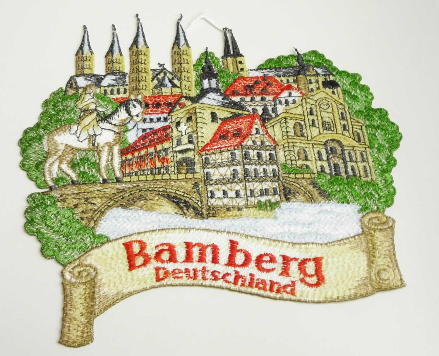 Stickerei, sog. Fensterbild aus Spinnstoff, Bamberg Deutschland, Foto siehe Anlage,
- stellt die Silhouette eine Stadt dar,
- in den Abmessungen von ca. 20 cm x 18 cm,
- laut Untersuchungsergebnis:
-- maschinelle Ätzstickerei als Motiv mit Stickfäden aus laut Antrag 100 % Polyester (synthetische Chemiefasern),
- mit einer aus der Stickerei herausgearbeiteten Aufhängevorrichtung,
- laut Antrag mit einem Wert von mehr als 35 EUR je kg Eigengewicht,
- ohne Hinweise auf Handarbeit.

"Ätzstickerei als Motiv, mit einem Wert von mehr als 35 EUR je kg Eigengewicht, nicht handgearbeitet"