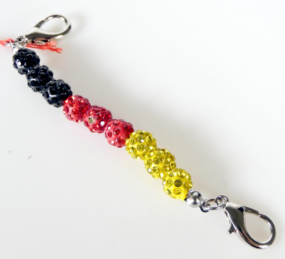 Es handelt sich um etwa 11 cm lange Zierelemente bestehend aus neun, mit Kunststoffsteinchen versehenen, ca. 0﻿,﻿8 cm großen Kugeln aus Kunststoff in den Farben schwarz, rot und gold (jeweils drei), die auf elastischen Monofilen aus Kunststoff kettenförmig aufgefädelt sind. An beiden Enden befinden sich Karabinerhaken aus unedlem Metall. Die Ware dient als Schmuckelement für Schuhe oder Taschen und ist zu jeweils zwei Stück gemeinsam verpackt.
- Antragstellerangaben -

Abbildung siehe Anlage.

Derartige Erzeugnisse gehören als "Fantasieschmuck, anderer als aus unedlen Metallen" zum TARIC-Code 7117 9000 00.