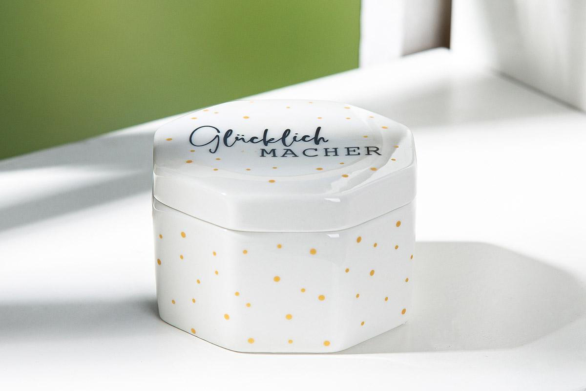 Anderer keramischer Ziergegenstand, sog. Dose, Foto siehe Anlage,
- achteckige, weiße, glasierte Dose mit einem lose aufliegenden, glasierten Deckel,
- auf der Außenseite gepunktet und auf dem Deckel mit einem Schriftzug "Glücklich MACHER" verziert,
- in den Abmessungen: ca. 8 cm (Breite) x 8 cm (Länge) x 5,3 (Höhe),
- keramisches Erzeugnis, nach der Formgebung gebrannt, tonmineralhaltiges Material enthaltend,
- lt. Antrag aus Porzellan,
- ohne Hinweise auf Handarbeit,
- stellt sich u. a. aufgrund der Ausstattung (Schriftzug), des äußeren Erscheinungsbildes (geringe Größe)
  als Ware dar, wie sie zum Ausschmücken oder Ausstatten von Wohnungen dient (z. B. als Bonbonniere
  oder als Schmuckdose),
- in einem Karton verpackt.

"Anderer keramischer Ziergegenstand, aus Porzellan, nicht handgearbeitet"
