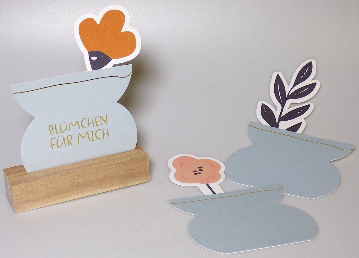 Es handelt sich um eine zusammengesetzte Ware, bestehend aus drei konturierten Zuschnitten aus Papier oder Pappe, jeweils in Form einer Blume oder eines Blätterzweiges in einer Vase, und einem Holzhalter mit Schlitz zum Aufnehmen/Aufstellen der Papier-/Pappzuschnitte.  Alle drei Zuschnitte sind einseitig farbig bedruckt und ein Zuschnitt weist zusätzlich den Schriftzug "Blümchen für dich" auf. Das Erzeugnis soll zu Dekorationszwecken verwendet werden.

Abildung siehe Anlage.

Derartige Waren gehören als "andere Bilddrucke" zum TARIC-Code 4911 91 00 90.