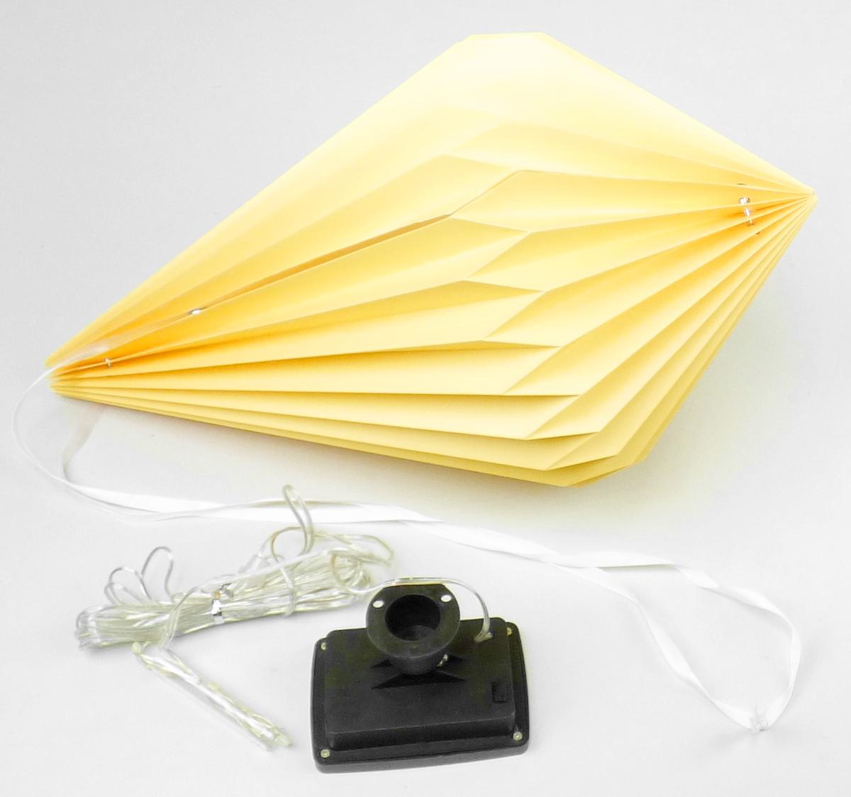 Bei dem sog. Solar Lampion Raute, Artikelnummern 9988183 und 9987782 handelt es sich um einen noch nicht zusammengesetzten elektrischen Beleuchtungskörper in Form eines faltbaren Lampions, bestehend aus einem rautenförmigen Gehäuse (d: ca. 300 mm, L: ca. 400 mm). Das Gehäuse ist aus gelbem lichtdurchlässigem Papier. Zur Stabilisierung ist der Lampion mit zwei Metallringen und Magnetkontakten ausgestattet. Im Innenraum des Gehäuses ist eine Lichterkette mit 10 LED-Lichtquellen eingesetzt, die mit einem Solarmodul (IP44) mit einem Ein- und Ausschalter verbunden ist. Das Erzeugnis kann mittels eines weißen Bandes aufgehängt werden. 
Der Lampion ist nicht handgefertigt.

Das Erzeugnis ist als "elektrischer Beleuchtungskörper, anderweit weder genannt noch inbegriffen, nicht von den Unterpositionen (HS) 9405 11 bis 9405 39 erfasst, photovoltaisch, ausschließlich zur Verwendung mit LED-Lichtquellen bestimmt, kein Scheinwerfer, aus Papier und Metall, nicht handgearbeitet" einzureihen.