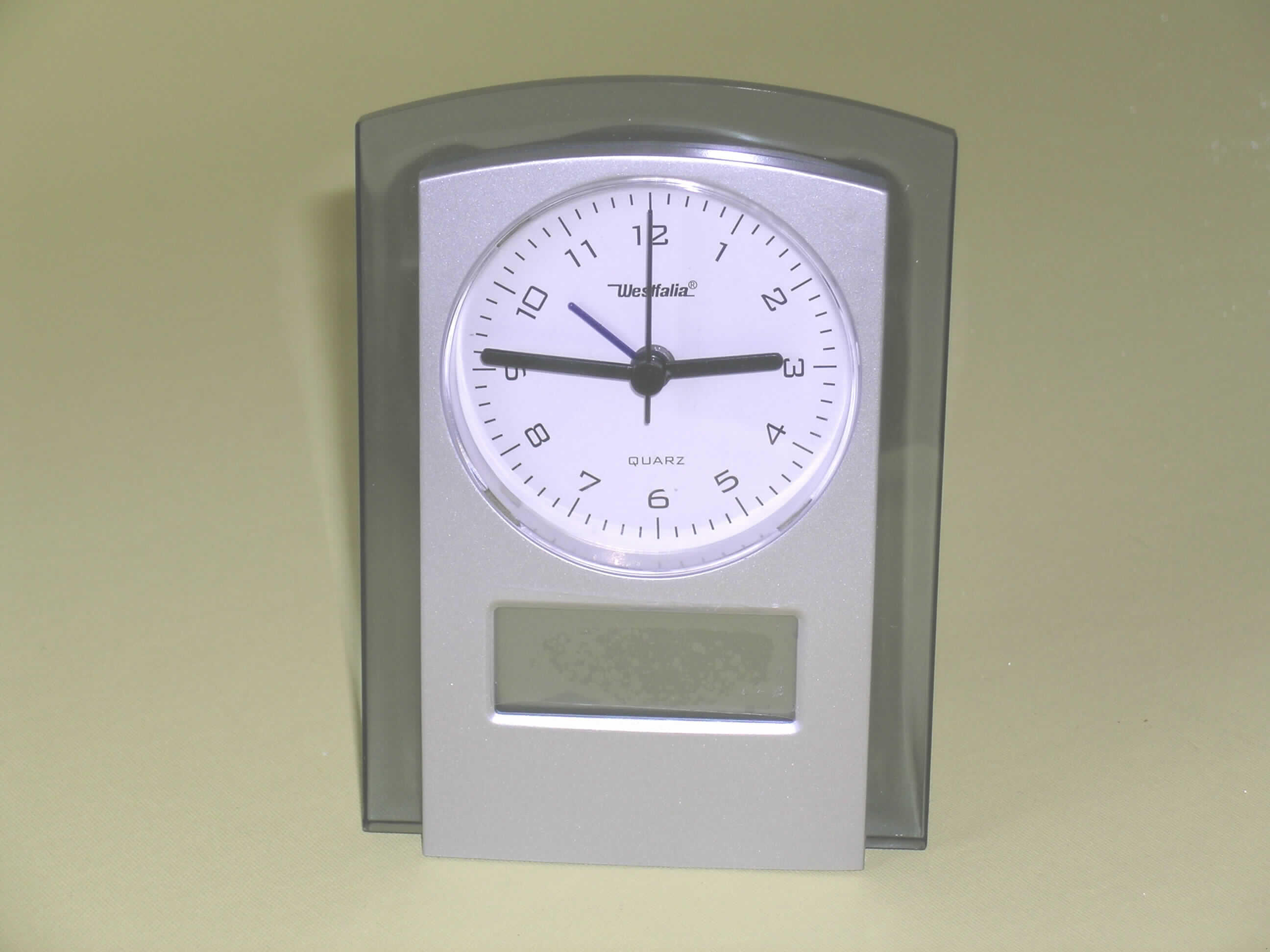 Andere als in den Positionen 9101 bis 9104 erfasste Uhr, Wecker, elektrisch betrieben (1,5 V-Batterie), in Form einer zusammengesetzten Ware, sog. Analog- und Digitaluhr mit Innenthermometer (Artikel-Nr.: 657718). Das Uhrwerk (kein Kleinuhr-Werk) des im Hinblick auf die Verwendung charakterbestimmenden Tisch-Weckers (ca. 14 cm hoch und 10 cm breit) befindet sich zusammen mit einem Innenthermometer mit Anzeige in Grad Celsius und Grad Fahrenheit auf einer gemeinsamen Platine in einem silberfarbenen Kunststoffgehäuse mit einem an drei Seiten umlaufenden durchsichtigen Kunststoffrahmen.

Unterhalb der analogen Uhr mit Stunden-, Minuten- und Sekundenzeiger sowie Zeiger für die Weckzeit befindet sich ein LCD-Feld für die Digitalanzeige von Wochentag, Datum und Temperatur. Auf der Rückseite des Weckers befinden sich der Uhrzeitstellknopf, der Weckzeitstellknopf, der Weckschalter (Ein/Aus), ein Knopf für Schlummerfunktion und Beleuchtung, drei Funktionstasten und ein Schiebeschalter für das Ablesen der alternativen Digitalzeit. 

Die Ware ist in einer gepolsterten Kunststofftüte zusammen mit einer Bedienungsanleitung in einem Pappkarton verpackt.