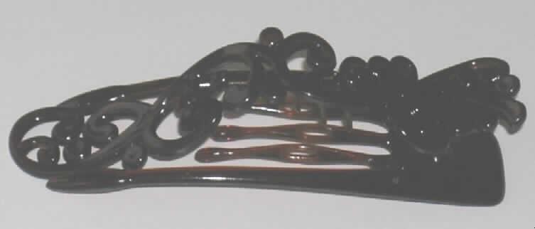 Haarklammer aus Kunststoff.
Es handelt sich um eine ca. 11 cm x 3,5 cm x 2,3 cm (Länge x Breite x Höhe) große Haarklammer aus braunem, transparentem Kunststoff. Die beiden Klammerteile sind durch eine Metallfeder miteinander verbunden. Der obere Klammerteil ist gebogen gearbeitet, weist ornamentartige Durchbrüche auf und ist auf der Unterseite mit drei Zähnchen ausgestattet. Der untere Klammerteil ist in Art eines Kammes mit zwei langen, äußeren und drei kürzeren innen angeordneten Zinken gearbeitet.
Das Erzeugnis dient zum Festhalten der Haare.
Die Haarklammer ist auf einer Kunststoffkarte mit Aufhängeöse festgesteckt.
