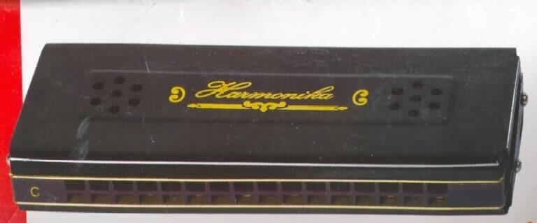 Blasinstrument in Form einer Mundharmonika, Länge 130 mm, bestehend aus einem Kanzellenkörper aus Stahlblech, mit zwei Reihen zu jeweils 16 Blasöffnungen (Kanzellen) für 16 Töne. Die Mundharmonika, die beiseitig bespielbar ist, ist in einer bedruckten Pappschachtel verpackt.