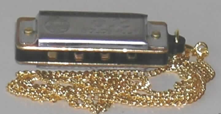 Blasinstrument in Form einer Mundharmonika, sog. Mini-Mundharmonika, Länge 40 mm, bestehend aus einem Kanzellenkörper aus Kunststoff und Stahlblecheinfassungen, mit 4 Blasöffnungen (Kanzellen) für 8 Töne (komplette Tonleiter). Die Mundharmonika, charakterbestimmend in Bezug auf die Verwendung, ist an einer etwa 58 cm langen goldfarbenen Kette aus unedlem Metall befestigt und in einer bedruckten Pappschachtel verpackt.