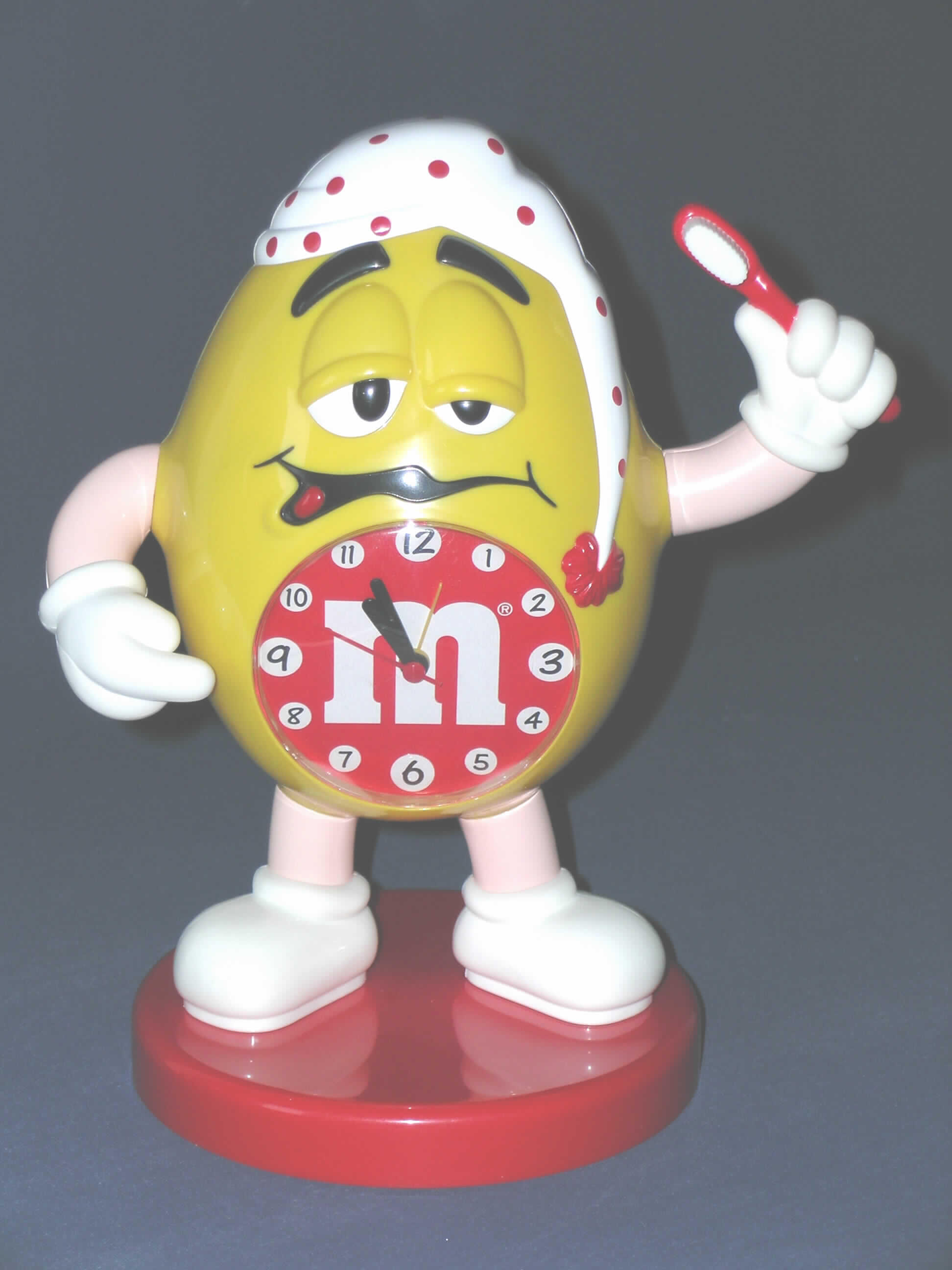Andere als in den Positionen 9101 bis 9104 erfasste Uhr, Wecker, elektrisch betrieben (1,5 V Batterie), sog. "M & M Schlafmann". Die Uhr ist in eine auf einem runden Sockel stehende M & M-Figur eingebettet (Gesamtmaße: ca. 26 cm hoch, 22 cm breit und 15 cm tief); Figur und Sockel bestehen aus Kunststoff.
Das Uhrwerk, dessen Zeitteilung durch einen Schwingquarz erfolgt, ist ca. 5,5 cm lang, 5 cm breit und 1,5 cm breit (kein Kleinuhrwerk). Uhrzeit und Weckzeit werden über zwei Drehknöpfe auf der Rückseite der Figur eingestellt und sind über eine Analoganzeige ablesbar. Zum Ein- und Ausschalten der Weckfunktion dient der linke Arm der Figur, die hier eine Zahnbürste in der Hand hält (Hand oben = Ein, Hand unten = Aus). 
Die Ware ist in einer durchsichtigen Kunststofftüte verpackt.