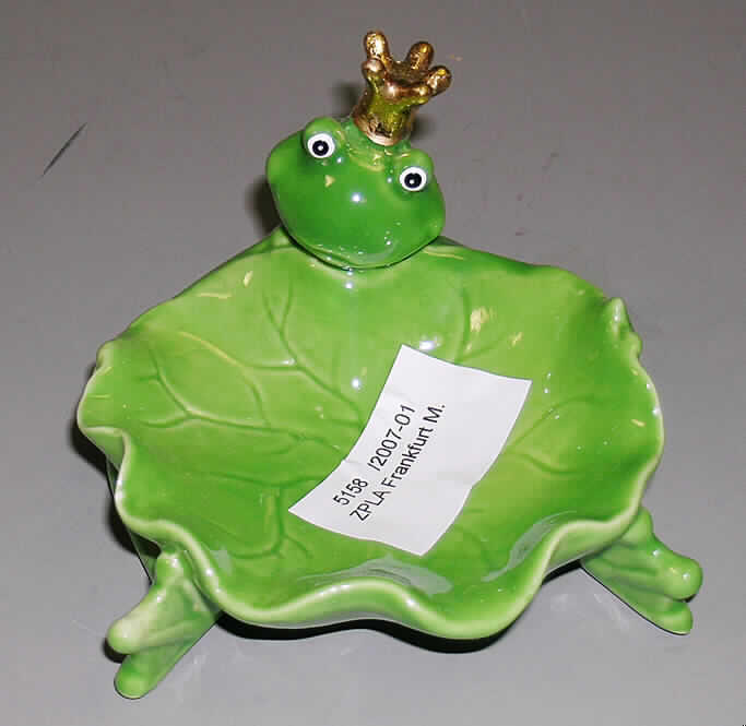 Toilettengegenstand aus Porzellan, sog. Schale "Froschkönig", Art. 24410, siehe Foto, 
- keramisches Erzeugnis, nach vorheriger Formgebung gebrannt, tonmineralhaltiges Material,
- nach augenscheinlicher Untersuchung handelt es sich um Porzellan;
  weißer, verglaster Scherben, durchscheinend, nicht ritzbar,
- überwiegend glasierte, grün bemalte Seifenschale in Form eines stilisierten Blatts,
- mit einem leicht hochgezogenen, wellenförmig gearbeiteten Rand,
- mit einer angearbeiteten, grün bemalten Nachbildung eines sitzenden Froschs mit 
  goldener Krone, der mit seinen Armen und Beinen die Seifenschale umschließt,
- mit einer Größe von ca. 11 x 12 x 9 cm, 
- die Verwendung als Seifenschale wird durch die Verzierung (angearbeiteter Frosch) 
  nicht eingeschränkt, somit keine Zuweisung als Ziergegenstand zu Position 6913.