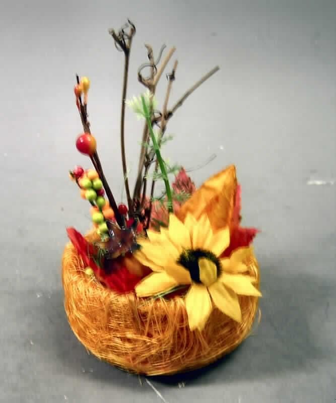 Ware aus künstlichen Früchten/Blattwerk/Blumen (lt. Antrag: "kl. Herbstgesteck im Sisalkörbchen",
siehe Abbildung), bestehend aus:
- einer Unterlage (Körbchen; ca. 13 cm hoch; mit einem Durchmesser von ca. 7 cm) aus pflanzlichen
  Flechtstoffen,
- auf der Oberseite vollständig verziert mit durch Zusammenfügen und Kleben in Blumenbindetechnik
  hergestellten künstlichen Nachbildungen von:
-- einer künstlicher Physalis aus überwiegend Papier,
-- einer künstlichen Blume aus überwiegend Spinnstoff,
-- künstlichen Beeren aus überwiegend Kunststoff,
-- zwei künstlichen Blättern aus Spinnstoff, in einem Stück hergestellt (gestanzt),
-- natürlichem pflanzlichem Material und
-- einem pflanzlichen Zweig aus Kunststoff, in einem Stück hergestellt, 
- im Hinblick auf den Umfang und das optische Erscheinungsbild (Zierwirkung) bestimmen die künst-
  lichen Früchte und Blumen den Charakter der Ware; bei diesen wiederum ist nach Umfang und Zier-
  wirkung Papier/Spinnstoff charakterbestimmend,
- für den Einzelverkauf in einer klarsichtigen Kunststofftüte aufgemacht.