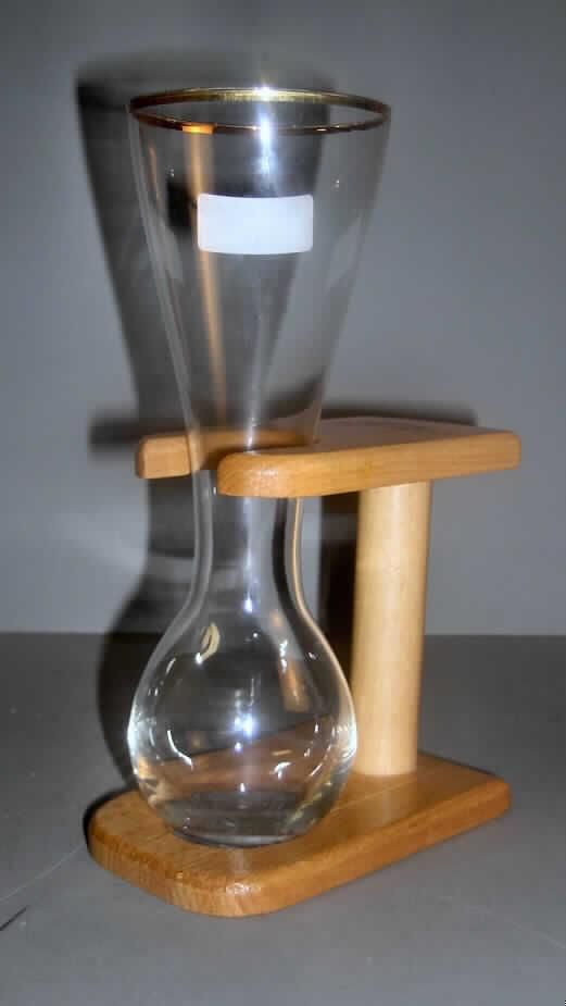 Trinkglas, Glasware zur Verwendung bei Tisch, sog. Kutscherglas, siehe Foto,
aus verschiedenen Bestandteilen zusammengesetzte Ware, bestehend aus
- einem Trinkglas ohne Stiel,
-- aus gewöhnlichem Glas, lt. Antragsangaben handgefertigt (manuelle Glasentnahme),
-- mit einem bauchigen Unterteil, zum oberen Ende hin trichterförmig gearbeitet,
-- entlang dem oberen Rand goldfarben bemalt, somit anders (weitergehend) bearbeitet,
-- mit einer Höhe von ca. 22 cm und einem Durchmesser (oberer Rand) von ca. 8 cm,
- einem Gestell aus Holz (Holzware zur Verwendung bei Tisch),
-- aus lackiertem Buchenholz (lt. Antragsangaben),
-- mit einer Standplatte mit einer runden Aussparung für das Unterteil des Trinkglases,
-- mit einem vertikal verlaufenden Stab als Verbindung der Standplatte mit der Halterung,
-- an der Halterung mit einer runden, zur Schmalseite offenen Aussparung für das Trinkglas,
-- mit einer Höhe von ca. 13 cm,
-- dient dem Einstellen des Trinkglases,
- das Trinkglas bestimmt insbesondere aufgrund seiner Bedeutung in Bezug auf die 
  Verwendung den Charakter der zusammengesetzten Ware.