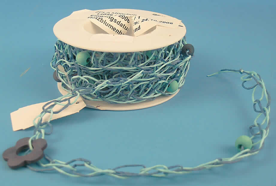 Häkelgalonborte (lt. Antrag: "Holzblumenband", siehe Foto)
- als Meterware,
- mit einer Breite von ca. 1,5 cm,
- ohne Flor- oder Schlingenoberfläche,
- ohne Elastomergarne oder Kautschukfäden,
- in Kettenwirktechnik hergestellt (Maschenstäbchenreihen),
- aus Papiergarnen (gedrehte Papierstreifen),
- mit eingearbeiteten kleinen Kugeln und Blüten aus Holz,
- für den Einzelverkauf auf einer Kunststoffrolle aufgemacht