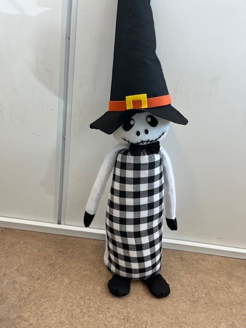 Varen er en halloweenfigur af tekstil med små fødder, en lang krop, lange arme, et hoved med et kranie-agtigt udtryk med aflange øjne, næsebor og en lang, sammensyet mund. Figuren bærer en høj, spids hat og en lille butterfly.
Varen er til dekoration ved højtiden halloween.
Varens fødder, bagside af kroppen, arme og hat er af filt, hovedet er af fleece, og forsiden af kroppen er af vævet tekstil. Det er oplyst, at varen er fyldt med sand og vat.
Varens fødder er sorte, kroppen er sort/hvid ternet på forsiden og hvid på bagsiden, armene er hvide med sorte hænder, hovedet er hvidt med sorte øjne, næsebor og mund, hatten er sort med et orange bånd rundt om. På det orange bånd er en gul firkant med hul i midten, som ligner et spænde.
Varen måler ca. 100 cm fra bunden til spidsen af hatten.