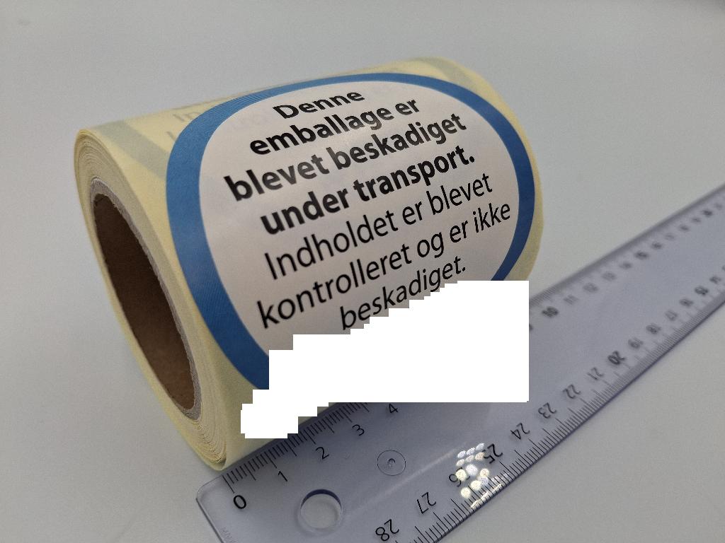 Varen er runde, trykte klistermærker af papir på et underlag af papir. Varen har en smal, blå ring langs kanten, et påtrykt firmalogo nederst og en påtrykt sort tekst på hvid baggrund i midten. Teksten fortæller, at selvom emballagen er beskadiget, er varen ikke beskadiget.
Varen anvendes til at gøre opmærksom på, at en vare er blevet kontrolleret og ikke er beskadiget.
Det er oplyst, at varen er af hvidt, træfrit papir, og at underlaget er af permanent glasinpapir.
Varen foreligger oprullet med 200 stk. pr. rulle. 
Varen har en diameter på 78 mm og foreligger på et underlag med en bredde på 8 cm.