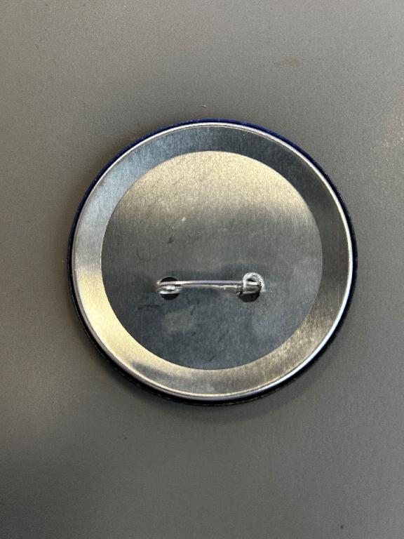 Varen er et rundt skilt med blålige og sorte farver med tekst og logo på forsiden. Bagsiden er af metal, og der sidder en nål til fastgørelse, som er fastgjort gennem to huller.
Varen er beregnet til fastgørelse på beklædning, og det er oplyst at varen anvendes som reklame, og at den bæres af butikspersonale i forbindelse med en bestemt kampagne.
Det er oplyst, at varen er fremstillet af metal, og at forsiden er beklædt med papir, som er dækket af plastik.
Fremsendt vareprøve har en diameter på ca. 7,5 cm.