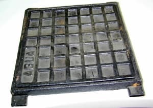Tapa de observación de fundición de hierro dúctil o maleable, obtenida por moldeo, que presenta un diseño en cuadrículas. La tapa es cuadrada de 18,5 cm de lado y el marco mide aproximadamente 19,8 cm de lado. En su parte superior izquierda viene escrita la palabra agua.