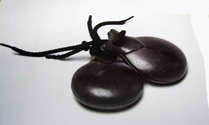 Instrumento musical de percusión, de madera, de color marrón, denominado castañuelas. El cordón que sujeta ambas piezas cóncavas es sintético. Se presenta para su venta al por menor en varios tamaños.