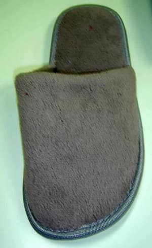 Calzado de casa. Zapatilla que deja el talón al aire, cubriendo casi todo el empeine. Parte superior de materia textil de color gris (poliéster 100%) y suela constituida por una plancha de plástico, que en su parte exterior, en contacto con el suelo, lleva una capa de materia textil muy fina de tejido de punto de fibras de poliéster (100%) que está adherida a la plancha plástica de la suela.