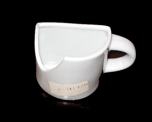 Artículo de cerámica de porcelana, sin decorar, que tiene forma de semicilindro hueco cerrado en la base, con asa. Se utiliza para colocar publicidad, en la superficie plana lateral. Tiene unas medidas aproximadas de 88 mm de alto y 89 mm de diámetro