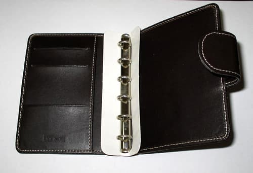 Cubierta de cuero para agenda. Dispone de una correa que cierra con un botón a presión y en el interior tiene un mecanismo de encuadernación de anilla para páginas interiores intercambiables y en una de sus tapas, incorpora un bolsillo y tres aberturas. Mide aproximadamente 126 x 100 x 22 mm.