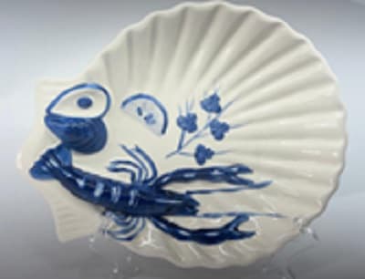 Descripción física: Articulo de uso doméstico de loza o barro fino consistente en bandeja de cerámica con forma de concha, de color blanco y con adornos en relieve, en color azul, con figuras de mariscos, crustáceos, rodajas de limón, etc. Esta perforado en el borde exterior con un agujero.
Medidas: 35 x 28 x 6 cm
Peso: 1,09 kg.
Función o uso: Según declaración del solicitante se utiliza como artículo de adorno, si bien, cumple todas las características para ser considerado un artículo de servicio de mesa o cocina, de uso doméstico.
Se presenta para su venta al público en una caja de cartón en formato de una unidad y con un soporte.