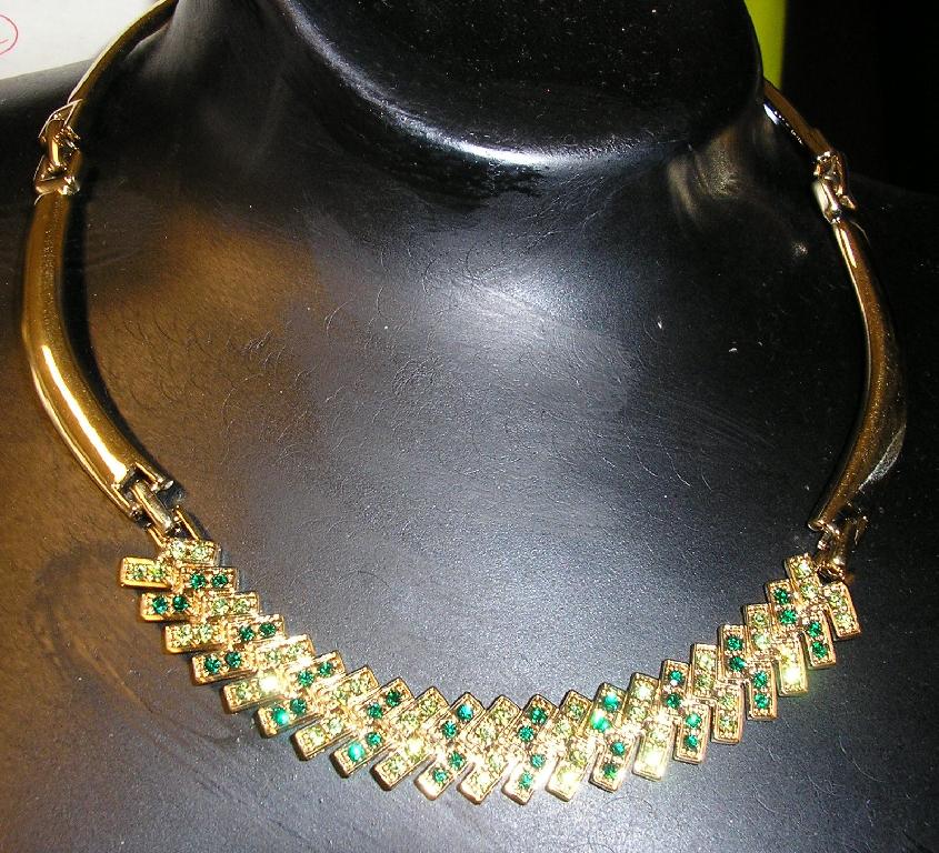 BIJOUTERIE DE FANTAISIE CONSISTANT EN UN COLLIER EN LAITON REVÊTU ÉLECTROLYTIQUEMENT D'OR (DE COULEUR JAUNE) SUR UNE SOUS-COUCHE DE NICKEL.

LE COLLIER EST RÉALISÉ À PARTIR DE CINQ PARTIES RIGIDES RELIÉES ENTRE ELLES PAR UN PETIT MAILLON ET IL SE FERME AU MOYEN D'UNE ATTACHE EN MÉTAL SUR LA NUQUE.

LA PARTIE CENTRALE DU COLLIER COMPORTE DES PETITS CRISTAUX DE VERRE (TEINTÉS), DE FORME RONDE, SERTIS SUR TOUTE SA LONGUEUR.