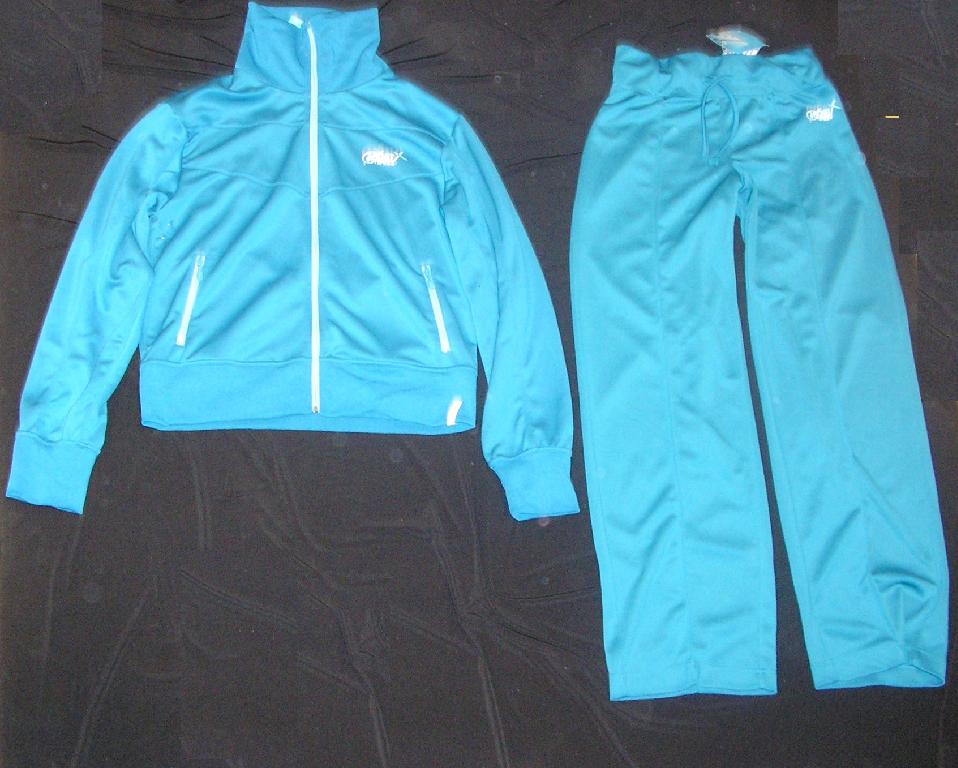 SURVÊTEMENT DE SPORT EN ÉTOFFE DE BONNETERIE DE FIBRES SYNTHÉTIQUES (100% POLYESTER), NON DOUBLÉ, COMPOSÉ DE DEUX PIÈCES : 

- UN GILET, TEINT UNIFORMÉMENT, DESCENDANT JUSQU'AU-DESSOUS DE LA TAILLE, AVEC UN COL MONTANT EN BORD-CÔTE, AVEC UNE OUVERTURE COMPLÈTE SUR LE DEVANT SE FERMANT À L'AIDE D'UNE FERMETURE À GLISSIÈRE, AVEC DES MANCHES LONGUES MUNIES D'UN BORD-CÔTE À LEUR EXTRÉMITÉ ET AVEC DEUX POCHES FENDUES LATÉRALES AVEC FERMETURE À GLISSIÈRE SITUÉES AU-DESSUS DE LA TAILLE. 
EN OUTRE, CE GILET PRÉSENTE UN LOGO IMPRIMÉ SUR LA POITRINE CÔTÉ GAUCHE, DEUX OEILLETS D'AÉRATION SOUS CHAQUE AISSELLE, UN LARGE BORD-CÔTE RAPPORTÉ À LA BASE ET COMPORTE DANS LE DOS UNE DÉCORATION RÉALISÉE À PARTIR D'UNE ENDUCTION PARTIELLE DE PETITS PICOTS DE MATIÈRE PLASTIQUE REPRÉSENTANT UN CORPS DE FEMME.

- UN PANTALON, TEINT UNIFORMÉMENT, DESCENDANT JUSQU'AUX CHEVILLES, DE COUPE LARGE, TENU À LA TAILLE PAR UNE LARGE BANDE ÉLASTIQUE ET UN CORDON COULISSANT, SANS OUVERTURE À LA TAILLE, NI POCHES.
IL PRÉSENTE UN LOGO IDENTIQUE À CELUI DU GILET IMPRIMÉ SUR LE HAUT DE LA JAMBE GAUCHE, DES NERVURES VERTICALES RÉALISÉES PAR SURPIQÛRE FORMANT DES PLIS SUR LE DEVANT ET LE DERRIÈRE DE CHAQUE JAMBE ET UN OURLET À L'EXTRÉMITÉ DE CHAQUE JAMBE.