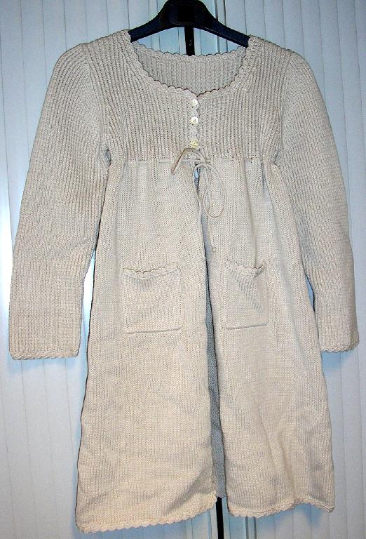 VÊTEMENT DE DESSUS POUR FILLETTES, DE TYPE MANTEAU, EN ÉTOFFE DE BONNETERIE DE COTON À GROSSES MAILLES, TEINTE UNIFORMÉMENT, NON DOUBLÉ, DESCENDANT JUSQU'AU-DESSUS DES GENOUX.

CE MANTEAU PRÉSENTE UNE ENCOLURE DE FORME ARRONDIE, UNE OUVERTURE COMPLÈTE SUR LE DEVANT SE FERMANT PARTIELLEMENT AU NIVEAU DE LA POITRINE, CÔTÉ DROIT SUR GAUCHE AU MOYEN DE TROIS BOUTONS, UN CORDON COULISSANT RESSERRANT À NOUER SOUS LA POITRINE, DES MANCHES LONGUES ET DEUX POCHES LATÉRALES PLAQUÉES SITUÉES SUR LES HANCHES.
LES MANCHES AINSI QUE LA PARTIE SUPÉRIEURE DU MANTEAU JUSQU'AU-DESSOUS DE LA POITRINE SONT CONFECTIONNÉES DANS UNE ÉTOFFE DE BONNETERIE TRICOTÉE DE TYPE INTERLOCK ALORS QUE LE RESTE DU VÊTEMENT EST EN BONNETERIE DE TYPE JERSEY.
LE POURTOUR DE L'ENCOLURE, DE L'OUVERTURE, DE L'EXTRÉMITÉ DES MANCHES, DES POCHES ET DE LA BASE EST BORDÉ PAR UN PETIT GALON TRICOTÉ DE FORME DENTELÉE.

POUR UNE TAILLE 8 ANS : 
- LONGUEUR DOS : ENVIRON 68 CM 
- POIDS : 420 GRAMMES