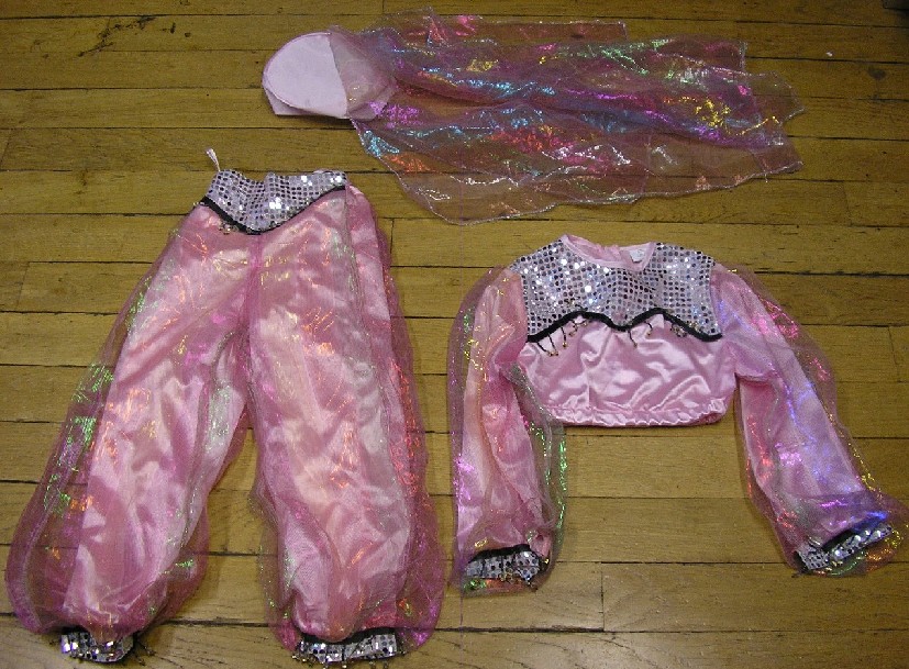 DÉGUISEMENT POUR ENFANTS EN ÉTOFFE DE BONNETERIE DE FIBRES SYNTHÉTIQUES (100% POLYESTER ) REPRÉSENTANT UNE DANSEUSE ORIENTALE.

CET ARTICLE COMPREND PLUSIEURS ÉLÉMENTS:

   - UN ENSEMBLE, RÉALISÉ DANS LA MÊME ÉTOFFE DE COULEUR UNIE, COMPOSÉ:
          - D'UN VÊTEMENT DESTINÉ À RECOUVRIR LA PARTIE SUPÉRIEURE DU CORPS DU GENRE BLOUSE COMPORTANT DES MANCHES LONGUES AVEC UNE OUVERTURE PARTIELLE À LEUR EXTRÉMITÉ FERMANT AU MOYEN D'UNE BANDE VELCRO, UNE ENCOLURE RONDE, UNE OUVERTURE PARTIELLE DANS LE DOS FERMÉE AU MOYEN D'UNE FERMETURE À GLISSIÈRE, 
          - D'UN PANTALON ÉLASTIQUÉ À LA TAILLE, DESCENDANT JUSQU'AUX CHEVILLES ET COMPORTANT UNE OUVERTURE PARTIELLE À LA BASE FERMANT AU MOYEN D'UNE BANDE VELCRO. 

CES DEUX VÊTEMENTS COMPORTENT DES EMPIÈCEMENTS DE TISSU TRANSPARENT (RAYONNE) AU NIVEAU DES MANCHES ET DES JAMBES, AINSI QUE DES PERLES SUR LE DEVANT ET À LEURS EXTRÉMITÉS.

   - UN CHAPEAU MUNI D'UN VOILE EN TISSU IDENTIQUE À CELUI DES EMPIÈCEMENTS DE L'ENSEMBLE.