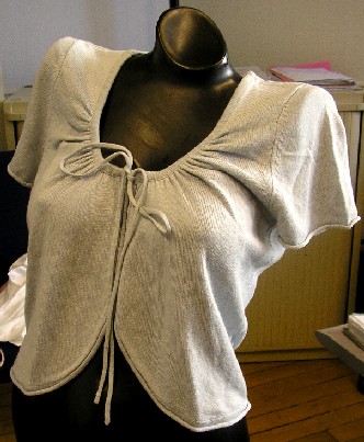 GILET COURT POUR FEMMES, DE TYPE BOLÉRO, CONFECTIONNÉ EN ÉTOFFE DE BONNETERIE MAJORITAIREMENT DE SOIE (85% SOIE - 15% CACHEMIRE), COMPORTANT MOINS DE 10 RANGÉES DE MAILLES PAR CENTIMÈTRE LINÉAIRE, UNICOLORE, RECOUVRANT LA POITRINE SANS ATTEINDRE LA TAILLE. 

CE VÊTEMENT PRÉSENTE UNE ENCOLURE ARRONDIE, UNE OUVERTURE COMPLÈTE SUR LE DEVANT SE FERMANT AU MOYEN DE DEUX LIENS COULISSANTS SITUÉS À L'ENCOLURE À NOUER SUR LE DEVANT ET DES MANCHES COURTES. 

LE POURTOUR DU VÊTEMENT (OUVERTURE, EXTRÉMITÉ DES MANCHES ET BASE) PRÉSENTE UNE FINITION ROULOTTÉE.