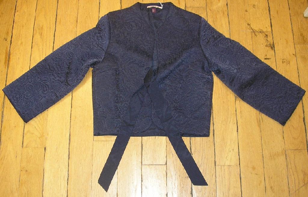 VESTE UNICOLORE POUR FEMMES EN TISSU MATELASSÉ DE SOIE, LES COUTURES FORMANT UN MOTIF FLORAL, DESTINÉE À RECOUVRIR LA PARTIE SUPÉRIEURE DU CORPS, DESCENDANT JUSQU'AUX HANCHES, AVEC 3 PANNEAUX COUSUS ENSEMBLE DANS LE SENS DE LA LONGUEUR, AVEC UNE ENCOLURE EN V SANS COL, AVEC DES MANCHES LONGUES POURVUES À LEURS EXTRÉMITÉS D'UN FIN GALON TON SUR TON, AVEC UNE OUVERTURE COMPLÈTE SUR LE DEVANT SE FERMANT AU MOYEN DE DEUX LIENS À NOUER, AVEC UNE DOUBLURE COMPLÈTE, AVEC UN GALON TON SUR TON SUR TOUT LE PORTOUR.