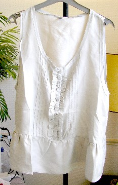 VETEMENT POUR FEMME, GENRE BLOUSE, DE FACTURE AMPLE, EN TISSU DE SOIE (100% SOIE), COUVRANT LA PARTIE SUPERIEURE DU CORPS, DESCENDANT JUSQU’AUX HANCHES, AVEC UNE ENCOLURE ARRONDIE DECOLLETEE DOTEE D’UN BIAIS SUR SON POURTOUR, AVEC UNE OUVERTURE PARTIELLE SUR LE DEVANT SE FERMANT DROITE SUR GAUCHE A L’AIDE DE BOUTONS. CETTE OUVERTURE PRESENTE DE CHAQUE COTE DES NERVURES ET UN VOLANT FORMANT JABOT, SANS MANCHES, AVEC UN BIAIS DE FINITION AUX EMMANCHURES, AVEC UN VOLANT A LA BASE, AVEC UN OURLET A LA BASE.