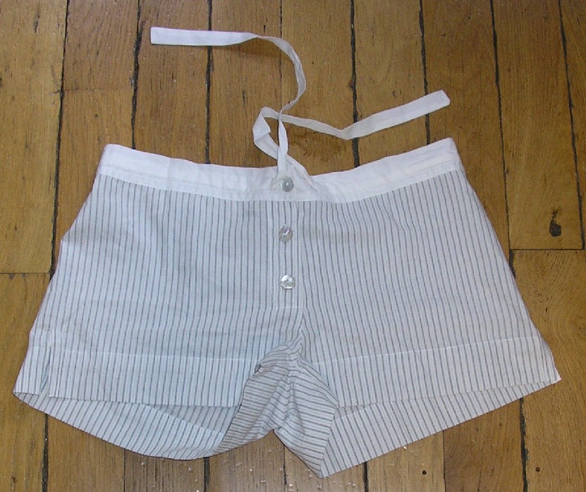 SHORT EN TISSU DE COTON, À RAYURES, DESCENDANT JUSQU'AU DESSUS DES CUISSES, AVEC UNE OUVERTURE SUR LE DEVANT SE FERMANT DROITE SUR GAUCHE AU MOYEN DE BOUTONS, AVEC UNE BANDE ÉLASTIQUE ET UN LIEN À NOUER AU NIVEAU DE LA TAILLE, AVEC DES FENTES SUR LES CÔTÉS, AVEC UN OURLET À LA BASE.