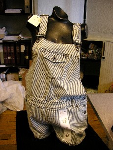 VETEMENT DESTINE A RECOUVRIR LA PARTIE INFERIEURE DU CORPS, GENRE SALOPETTE-SHORT, POUR FEMMES, EN TISSU 100% COTON, COMPORTANT UNE BAVETTE AVEC UNE POCHE A RABAT ET BOUTON AU NIVEAU DE LA POITRINE, DE LARGES BRETELLES REGLABLES SE CROISANT DANS LE DOS, UN SHORT SANS OUVERTURE, AVEC UNE POCHE EN BIAIS DE CHAQUE COTE SOUS LA TAILLE, UNE OUVERTURE PARTIELLE DE CHAQUE COTE DES HANCHES AVEC POUR FERMETURE DES BOUTONS, UNE POCHE AVEC RABAT ET BOUTON DE CHAQUE COTE DES FESSES, DES PASSANTS POUR CEINTURE AU NIVEAU DE LA TAILLE, LA BASE DES CUISSES EST OURLEE, DECORATION BANDE AUTO-ADHESIVE AVEC MOTIFS SUR DE DEVANT DE LA CUISSE GAUCHE.
