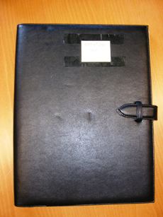 PORTE-DOCUMENTS DE TYPE CONFERENCIER, DE FORMAT 32 X 24 X 2 CM, A SURFACE EN FEUILLE DE MATIERE PLASTIQUE GRAINEE (POLYCHLORURE DE VINYLE) SE FERMANT SUR LA TRANCHE PAR UNE PATTE ET UN PASSANT. 
CET ARTICLE PRESENTE A L'INTERIEUR :  
- SUR LA PARTIE DROITE , UN RABAT DANS LEQUEL EST INSERE UN BLOC DE PAPIER AMOVIBLE, 
- AU CENTRE EN HAUT, UNE PASSANT ELASTIQUE POUR MAINTENIR UN STYLO, 
- SUR LA PARTIE GAUCHE, UNE GRANDE POCHE ET TROIS PETITES POUR RANGER DIVERS DOCUMENTS.