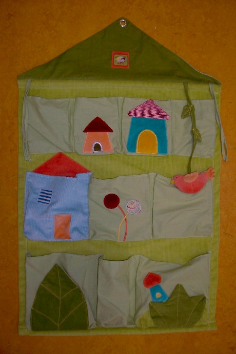 ARTICLE D’AMEUBLEMENT SE PRESENTANT SOUS LA FORME D’UN VIDE-POCHES, EN TISSU VELOURS COTELE DE COTON (100% COTON). IL EST DE FORME RECTANGULAIRE AVEC UN TRIANGLE SUR LE DESSUS FORMANT LA DEVANTURE D’UNE MAISON. IL COMPORTE UNE BAGUETTE EN BOIS GLISSEE A L’INTERIEUR DE LA BASE DU TRIANGLE. IL EST COMPOSE DE QUATRE PETITES POCHES VERS LE HAUT, DE TROIS GRANDES POCHES AU MILIEU ET DE DEUX GRANDES POCHES ET UNE PLUS PETITE VERS LE BAS.
CHAQUE POCHE EST DOTEE DE MOTIFS DECORATIFS EN VELOURS OU EN JERSEY FORMANT DES FLEURS, DES FEUILLES ET DES MAISONS.

CET ARTICLE COMPORTE SUR L’ENVERS DES BOUCLES ET ŒILLETS METALLIQUES PERMETTANT DE L’ACCROCHER AU MUR ET DES LIENS A NOUER A CHAQUE ANGLE PERMETTANT DE L’ATTACHER A UN LIT. IL EST DOTE D’UN ECUSSON.

CE VIDE-POCHES PEUT S'ADAPTER A DE NOMBREUX ENDROITS : LIT, MUR, TABLE A LANGER……….