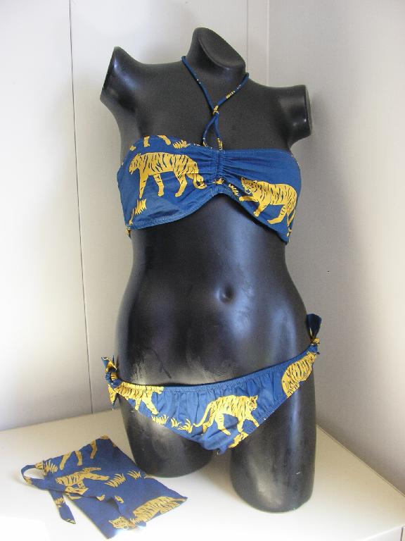 MAILLOT DE BAIN POUR FEMMES, CONFECTIONNÉ EN TISSU DE COTON (100%), COMPOSÉ :
- D'UN BANDEAU COMPORTANT UN LIEN RESSERRANT AU MILIEU DE LA POITRINE QUI PEUT S'ATTACHER AUTOUR DE COU. LE BANDEAU EST ÉGALEMENT SMOCKÉ SUR TOUT LE DOS, AVEC UN EFFET ÉLASTIQUÉ POUR ÊTRE MAINTENU AISEMENT.
- D'UN SLIP ÉCHANCRÉ COMPORTANT DES LIENS A NOUER DE CHAQUE COTÉ AU NIVEAU DES HANCHES ET D'UN ÉLASTIQUE SUR TOUTE LA BORDURE ET AU NIVEAU DE L'ENTREJAMBE. LE FOND EST DOUBLÉ ET DES FRONCES DÉCORATIVES SONT PRÉSENTES SOUS L'ÉLASTIQUE.

LE MAILLOT DE BAIN EST ACCOMPAGNÉ D'UNE POCHETTE CONFECTIONNÉE EN TISSU DE COTON (100%), DE FACTURE SIMPLE, SANS DOUBLURE INTÉRIEURE, DE FORME RECTANGULAIRE, AVEC UN LIEN RESSERRANT A NOUER EN HAUT DE L'ARTICLE EN GUISE DE FERMETURE.