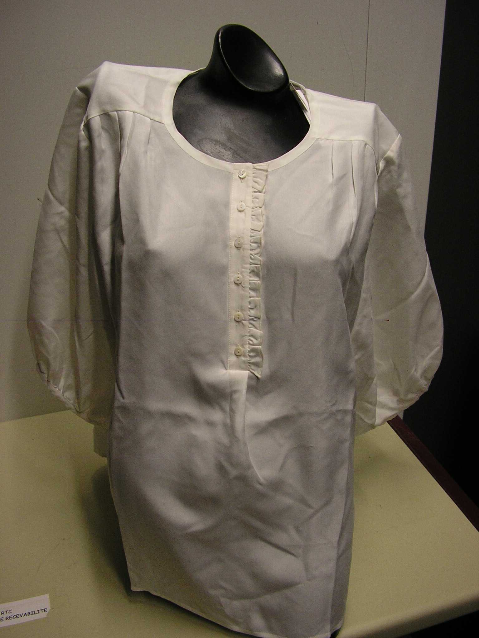 BLOUSE-CHEMISIER POUR FEMME, CONFECTIONNÉE EN SOIE, DE FACTURE FLUIDE ET LÉGÈRE, DESTINÉE À COUVRIR LA PARTIE SUPÉRIEURE DU CORPS ET DESCENDANT JUSQU'AUX HANCHES. CE VÊTEMENT PRÉSENTE UNE ENCOLURE ARRONDIE DOTÉE D’UN BIAIS SUR SON POURTOUR, UNE OUVERTURE PARTIELLE SUR LE DEVANT MUNIE DE VOLANTS SUR UN DES COTÉS ET SE FERMANT DROITE SUR GAUCHE AU MOYEN DE PETITS BOUTONS, DES MANCHES 3/4 DOTÉES D’UN BIAIS ET D'UN PETIT BOUTON A LEURS EXTRÉMITÉS, ET UN OURLET A LA BASE. LA BLOUSE-CHEMISIER EST ÉGALEMENT MUNIE DE PLIS DANS LE HAUT DU DOS ET AU NIVEAU DES ÉPAULES.