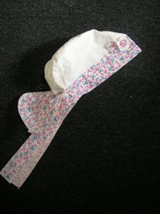 COIFFE COMPOSE DE TROIS BANDES DE TISSU UNI, AVEC UN BIAIS IMPRIME SUR LE POURTOUR SE TERMINANT PAR DEUX LANIERES FLOTTANTES AU DOS, AVEC UNE LANGUETTE DANS LE MEME TISSU POUR PROTEGER LA NUQUE, AVEC DEUX FLEURS EN TISSU COUSUES SUR LE DEVANT.  
COIFFE POUR JEUNES ENFANTS.