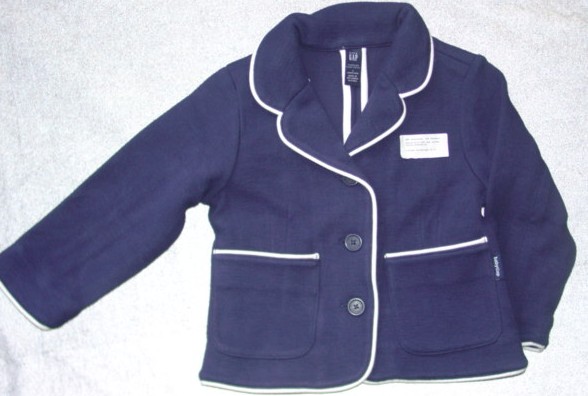 A KNITTED, RIBBED BLAZER STYLE JACKET FOR GIRLS. WITH A RIGHT OVER LEFT BUTTON CLOSURE. WITH PATCH POCKETS TO LOWER FRONTS. WITH LONG SLEEVES AND A REVER COLLAR. CONTRASTING PIPING TO ALL SEAMS AND COLLAR. 100% COTTON.
