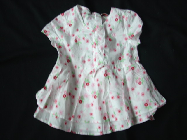 A WOVEN, PRINTED BLOUSE FOR GIRLS. REACHING DOWN TO BELOW THE WAIST. V-NECK. PARTIAL OPENING AT THE BACK FASTENED BY ZIP. WITH FALSE VERTICAL DRAWSTRING TO CENTRE FRONT, WITH DECORATIVE BOW AT WASIT. WITH CAPPED SLEEVES. HEMMED AT SLEEVE ENDS AND BASE OF GARMENT. THE GARMENT FLARES BELOW THE WAIST, AND HAS TWO LAYERS OF DIFERENT LENGTHS. 100% COTTON