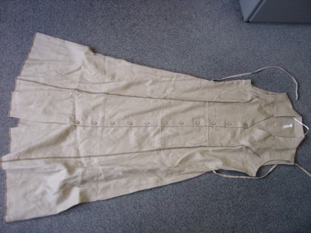 WOMANS WOVEN SLEEVELESS DRESS. REACHING DOWN TO BELOW THE KNEE. V NECK WITH COLLAR.  COMPLETE FRONT OPENING FASTENED RIGHT OVER LEFT BY BUTTONS.   HEMMED AT BASE. WITH TIES EXTENDING FROM SIDE OF WAIST. GARMENT IS MADE OF100 PERCENT LINEN.