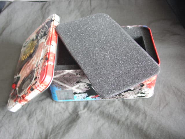A SMALL RETANGULAR STEEL BOX CONTAINER. TIN PLATED. WITH A WALL THICKNESS OF LESS THAN 0.5MM. CONTAINING PACKING MATERIAL IN THE FORM OF A CARDBOARD UNPRINTED PAPER PLATFORM AND FOAM SPONGE INSERT WITH CUT OUT SQUARE SECTIONS TO HOLD EVENTUAL CONTENTS IN PLACE.