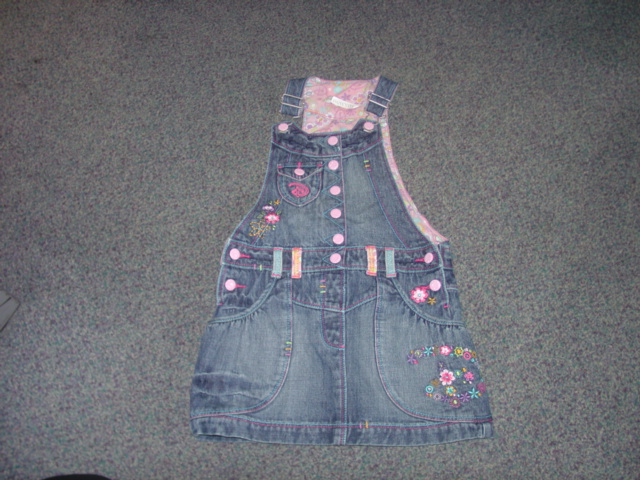 A WOVEN DENIM SKIRT FOR A CHILD. WITH BIB AND BRACE CHEST, DESIGNED TO COVER THE BODY FROM THE SHOULDERS TO MID THIGH. WITH BUCKLE CLIPS ON SHOULDER STRAPS. WITH POCKETS BELOW THE WAIST AND ONE ON THE CHEST. WITH EMBRIODERY AND BUTTON DETAIL. WITH 2 BUTTONS EACH SIDE OF THE WAIST. HEMMED. 100 PER CENT COTTON.