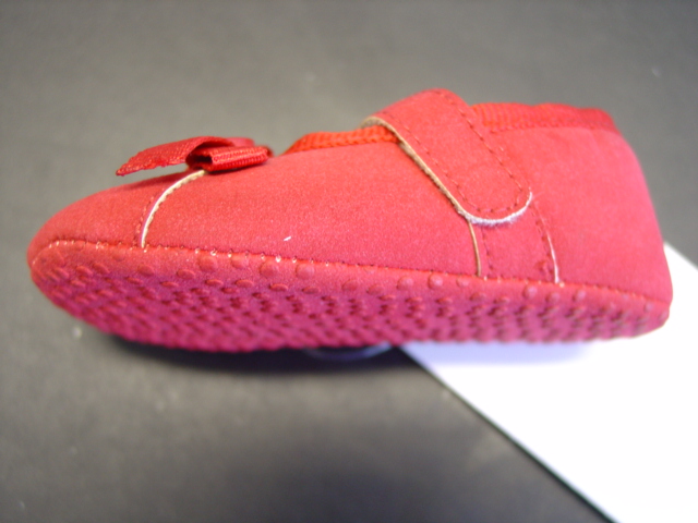 A SHOE DESIGNED FOR A BABY. THE UPPER IS MADE OF (IMITATION SUEDE) PLASTIC WITH AN OUTER SOLE OF PLASTIC WITH PLASTIC DOTS.  THE SHOE IS FASTENED WITH A SINGLE VELCRO STRAP. WITH DECORATIVE BUTTON BOW DETAIL. SHOE COLOUR IS RED.