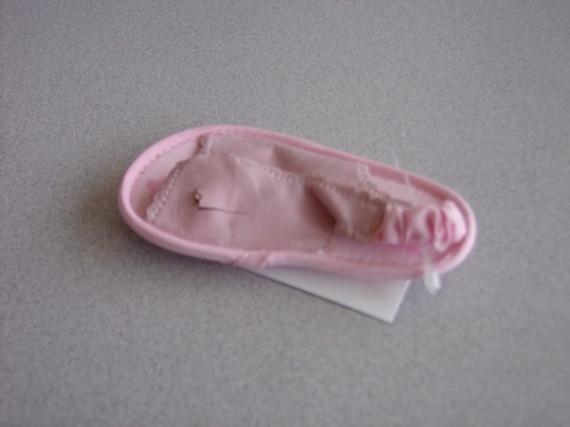 INDOOR FOOTWEAR FOR A BABY. NOT COVERING THE ANKLE.  UPPER OF A SINGLE PLASTIC STRAP ACROSS THE FRONT OF THE FOOT WITH A LOOP IN THE MIDDLE GIVING A SLIGHT RUCHED EFFECT . ELASTICATED TEXTILE  STRAP AROUND THE BACK OF THE HEEL. OUTER SOLE OF PLASTIC  WITH A FLOWER PATERN. SOLE EDGED WITH TEXTILE BINDING. PINK IN COLOUR