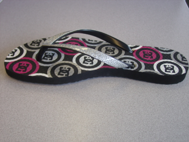 FLIP-FLOPS FOR WOMEN. THE UPPER STRAP IS MADE OF BLACK PLASTIC AND HAS A SILVER GLITTER SURFACE. STRAP IS FIXED TO THE SOLE BY MEANS OF PLUGS. THE SOLE IS OF RUBBER.